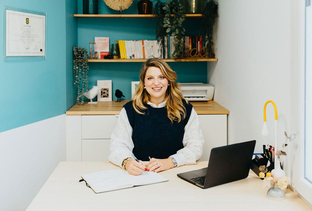 Clarisse Meynet, thérapeute, souriante dans son cabinet à Bordeaux, assise à son bureau avec un carnet et un ordinateur portable, entourée de livres et de décorations apaisantes.